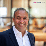 A man in a navy suit and white shirt smiles at the camera indoors. The background is blurred, with the Memcyco logo visible in the top right corner, highlighting Memcyco at Fraud Fight Club Round III.