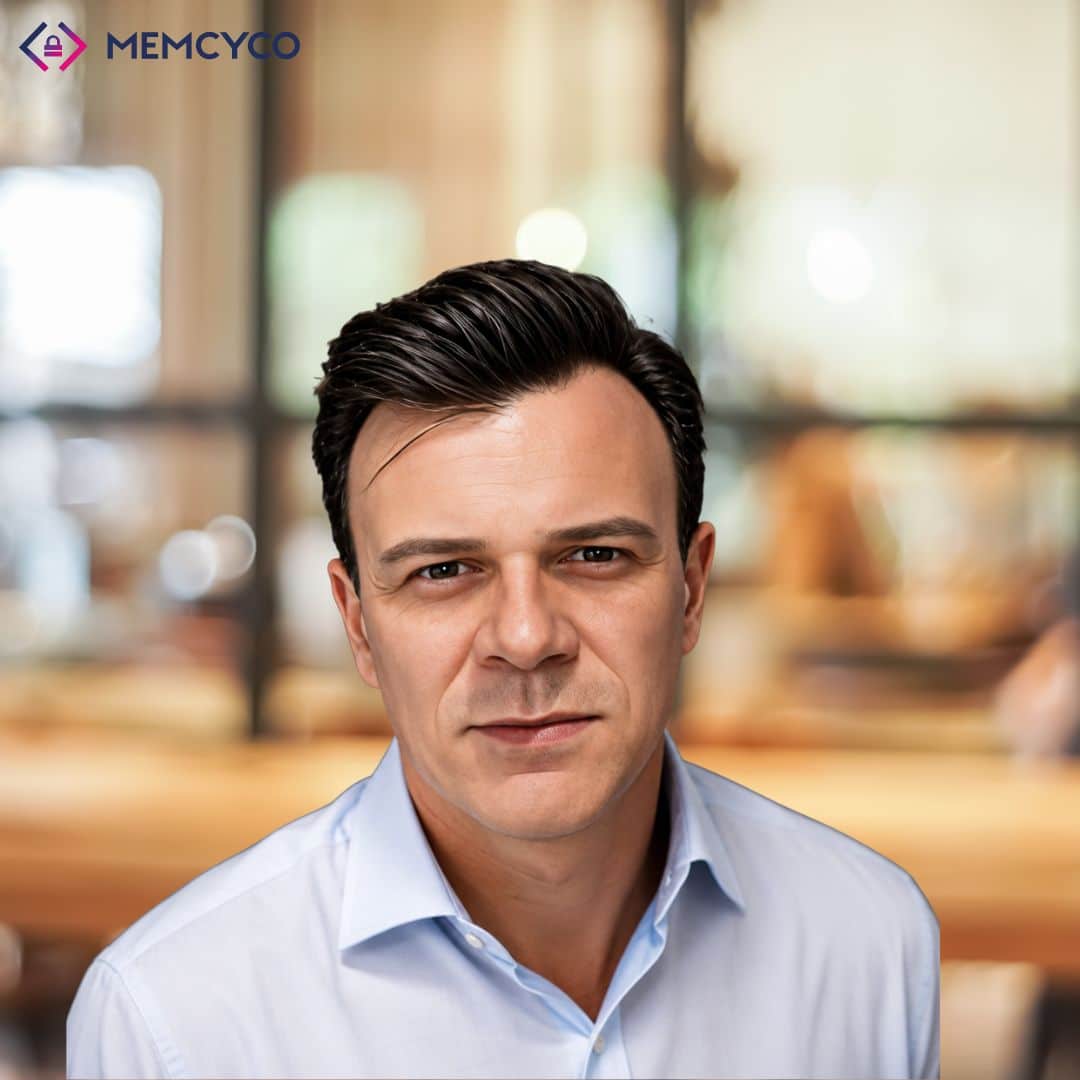A man with short dark hair wearing a light blue collared shirt sits indoors, with a blurred background of a modern office or cafe. The MEMCYCO logo appears in the top left corner, highlighting Memcyco at Global Anti-Scam Summit America 2026.